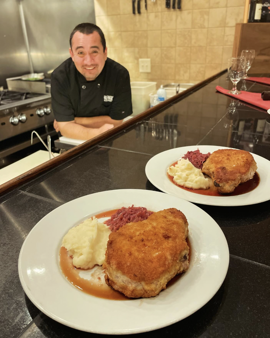 Chef souriant en veste noire derrière un comptoir avec deux assiettes de viande panée, purée de pommes de terre et chou rouge.
