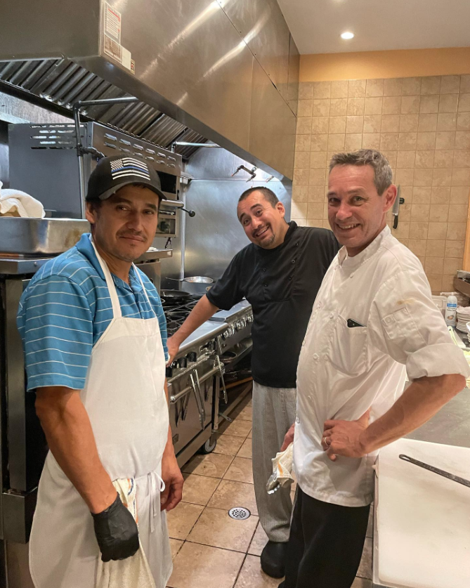 Trois cuisiniers souriants posant ensemble dans une cuisine professionnelle avec des équipements de cuisson en arrière-plan.