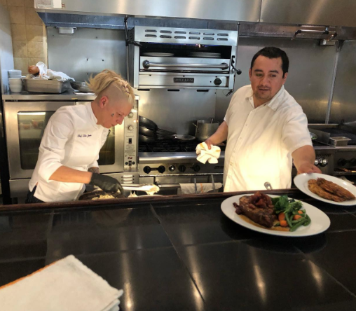 Deux chefs en cuisine, une femme préparant un plat et un homme servant des assiettes avec viande et légumes.